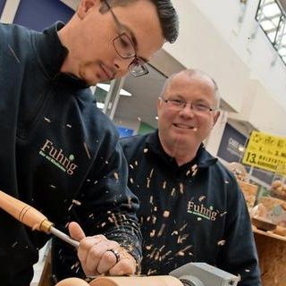 Peter Fuhrig Zwei Männer arbeiten gemeinsam an einem Holzprojekt, Späne fliegen umher.