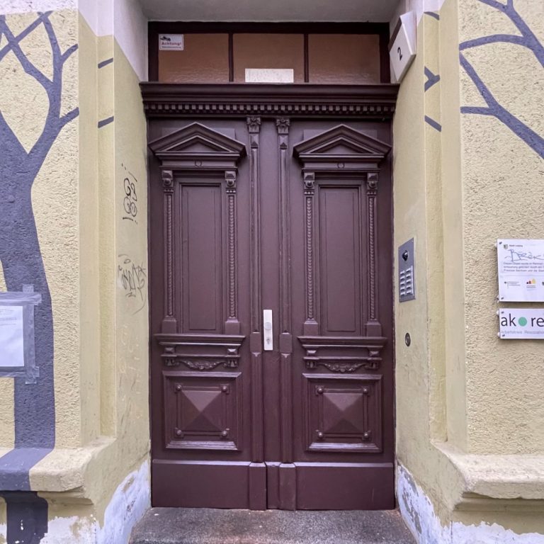 Braune Doppeltür mit dekorativen Elementen, umgeben von gelbem Putz und Baum-Motiven.