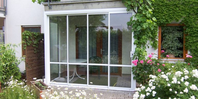 Veranda mit Glaswänden, umgeben von Blumen und Pflanzen.