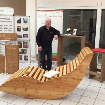 Karsten Fuhrig Ein Mann steht neben einer Holzliege in einem Ausstellungsraum.